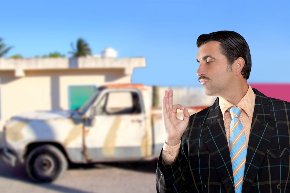 Image: a used-car salesman makes an OK sign with his fingers in front of a dilapidated pickup truck.