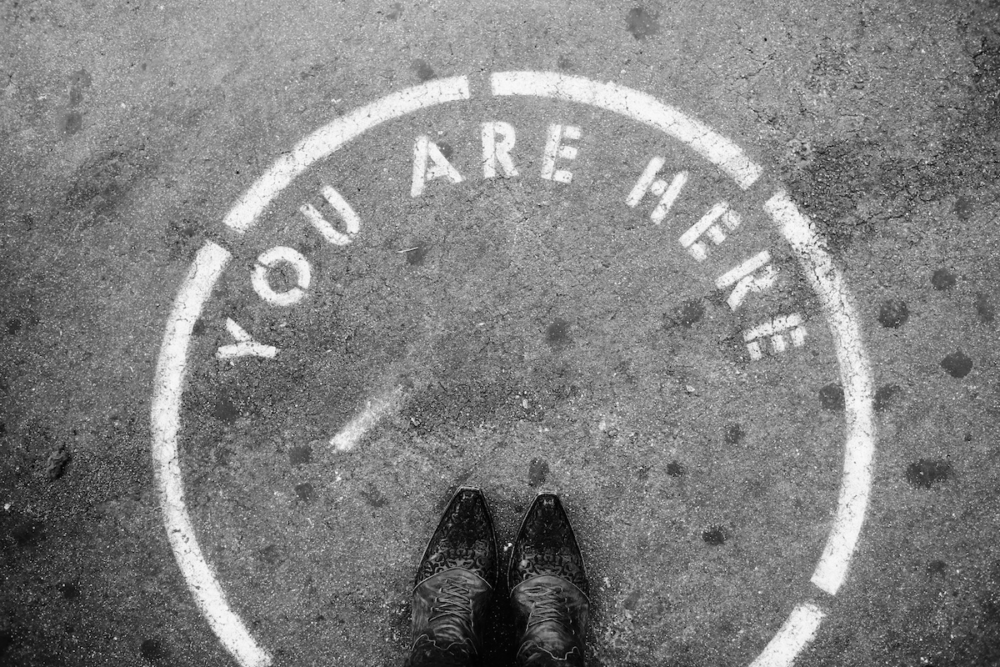 Image: the viewer looks down at a pair of shoes that are standing in a circle that's stenciled on the road along with the words "You are here."