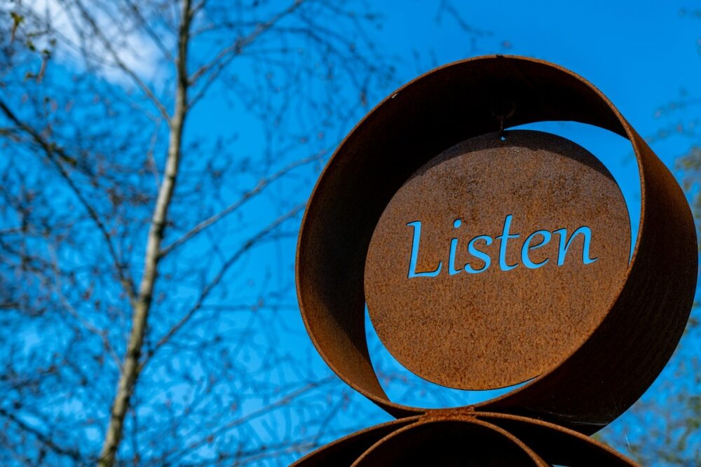 Image: at the Sculpture by the Lakes park in Dorchester UK, a rusted metal artwork has the word "Listen" cut out so that the deep blue sky shows through.