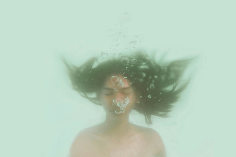 Image: a woman is underwater with her eyes closed, exhaling and releasing air bubbles that float upward.