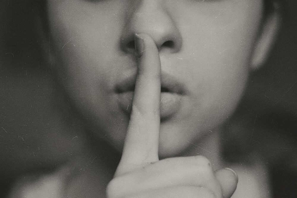 Image: a photo of a woman holding a finger in front of her lips in a silencing gesture.