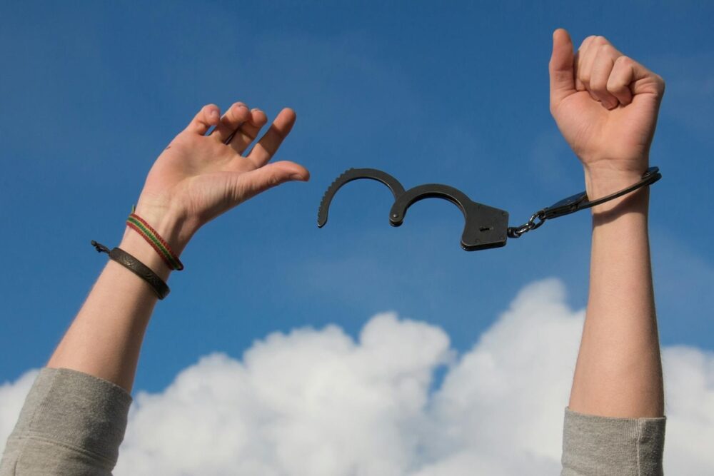 Image: a woman's arms are upraised against the blue sky. One half of a pair of handcuffs is secured to one wrist, while her other wrist has just broken free.