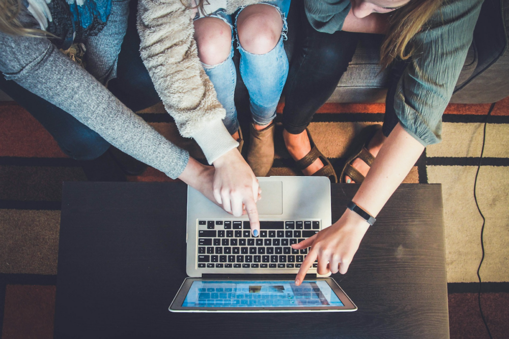 Image: the camera looks down at the hands of three women sitting together on a sofa, all pointing at the screen of an open laptop computer.