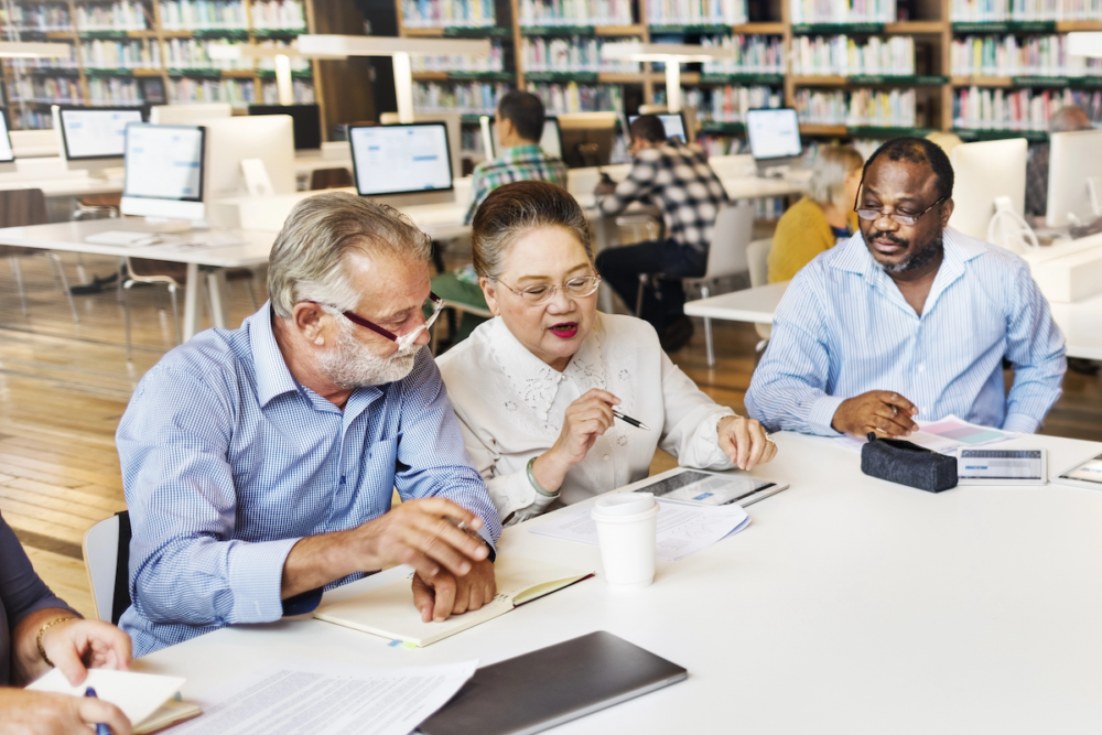 Image: a group of people are seated at a table in a library, having a discussion.