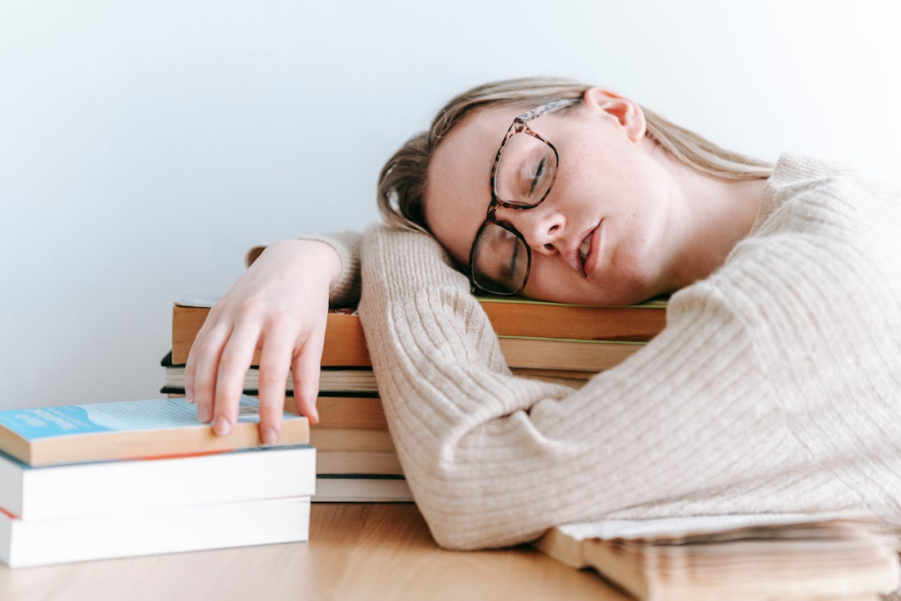Image: at a table, a woman has fallen asleep on a stack of books