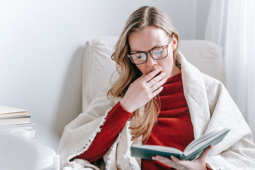 Image: a woman draped in a shawl and sitting in a chair yawns while trying to read a book.